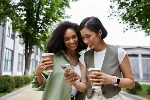 Two women, a lesbian couple, walk down a city street, smiling and looking at a phone together. — Stock Photo
