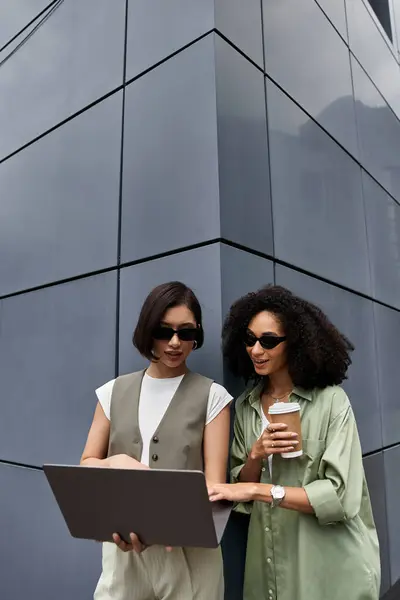 Two women in stylish outfits, look at a laptop outside a building. — Stock Photo