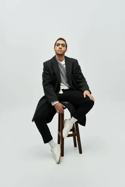 Un uomo afroamericano elegante posa in un cappotto alla moda, elegante e scarpe da ginnastica bianche mentre si siede su uno sgabello su uno sfondo grigio. — Foto stock
