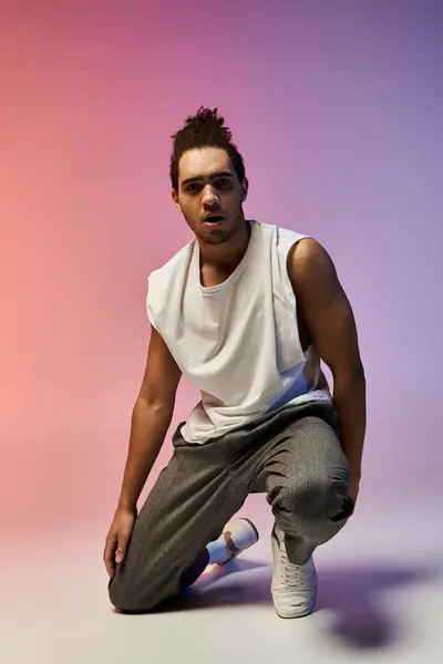 Un jeune Afro-Américain vêtu d'une chemise blanche sans manches et d'un pantalon gris pose sur un genou sur un fond dégradé lumineux et coloré. — Photo de stock