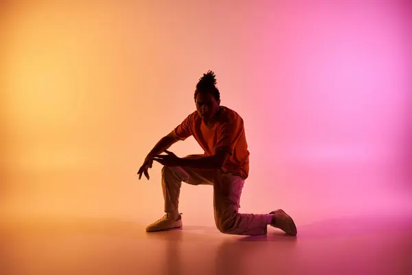 A young African American man in a vibrant orange shirt dances in a studio, illuminated by a gradient of pink and yellow lights. — Stock Photo