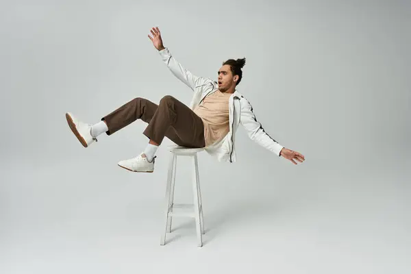 A young African American man in a stylish outfit poses dramatically on a grey background, sitting on a stool with one leg extended. — Stock Photo