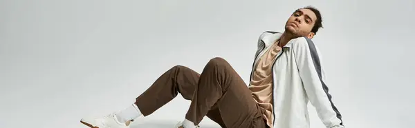 A young African American man in a stylish white jacket and brown pants poses against a grey background. — Stock Photo