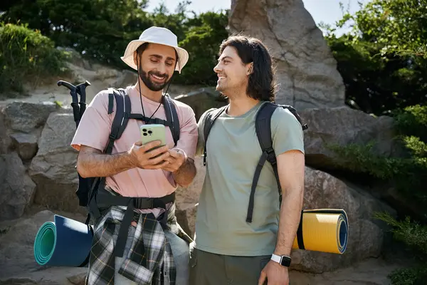 Two young men, a gay couple, enjoy a hiking trip through scenic wilderness. They are smiling and happy, looking at a phone. — Stock Photo