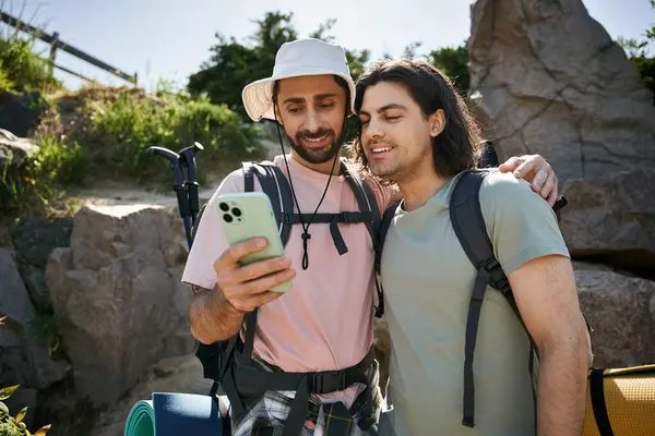 Um jovem casal gay gosta de uma caminhada de verão, olhando para seu telefone juntos enquanto vestindo mochilas. — Fotografia de Stock