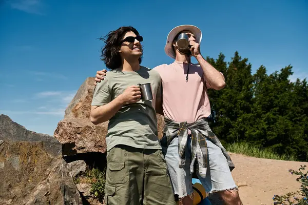 A young gay couple enjoys a summer hike in the wilderness, taking a break to sip from their mugs and admire the view. — Stock Photo