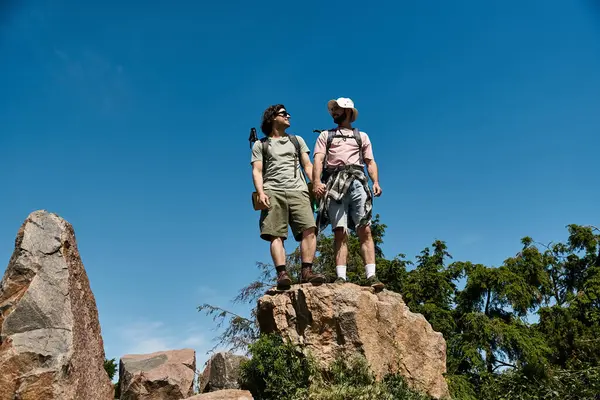 Two young men, a gay couple, stand hand-in-hand atop a rock formation on a mountain hike in the summer. — Stock Photo