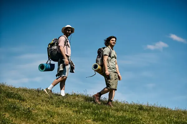 Dois jovens caminham em um campo ensolarado com mochilas, sorrindo em sua aventura ao ar livre. — Fotografia de Stock