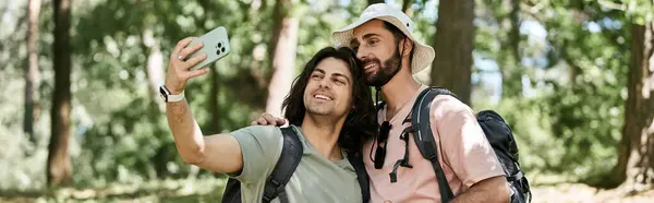 Two young men, a gay couple, hike through a forest on a sunny summer day. They take a selfie to capture the moment. — Stock Photo