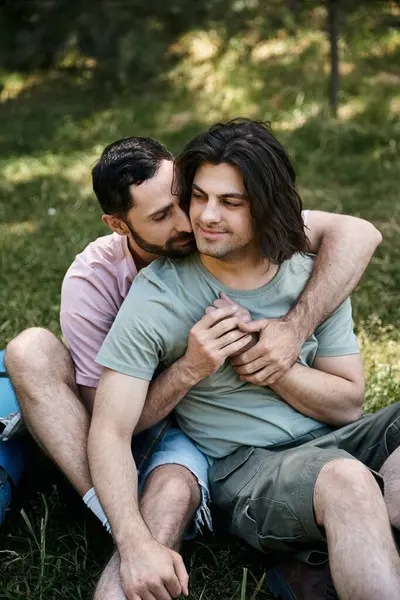 A young gay couple sits on a grassy hillside in a forest, embracing and showing affection. — Stock Photo