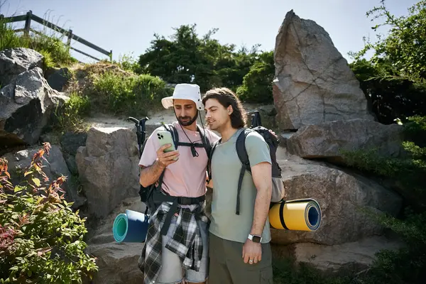 Two young men, a gay couple, are hiking in a rocky wilderness area on a sunny summer day. They are checking a map on a phone. — Stock Photo