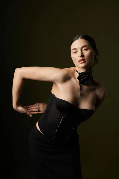 A young woman with brunette hair wears a black corset and a floral choker while posing against a black background. — Stock Photo