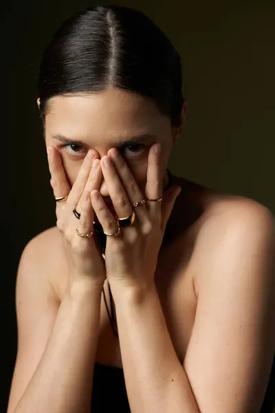 A young woman with brunette hair poses against a black background, her hands covering her face, revealing a subtle mystery. — Stock Photo