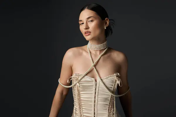 A young woman in a corset stands against a dark background, her neck adorned with a pearl necklace and her eyes looking away. — Stock Photo