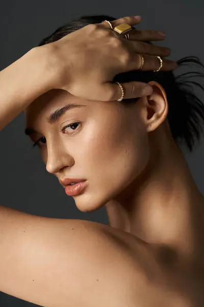 Young woman with dark hair posing against dark background, delicately resting hand on head, showcasing multiple golden rings on fingers. — Stock Photo