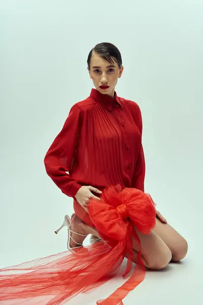 Young woman with short hair in a beautiful red dress with bow, sitting gracefully on a grey background. — Stock Photo