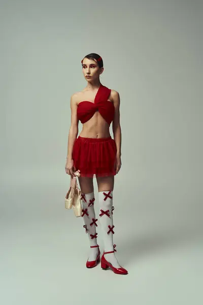 Young woman in red crop top, mini skirt poses on grey backdrop. Short hair, red bow, white socks create contrast. — Stock Photo