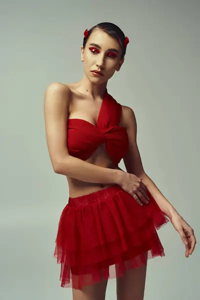 Una joven confiada en top rojo y mini falda irradia poder con un maquillaje audaz sobre un fondo gris. - foto de stock