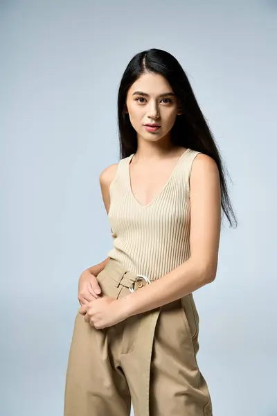 Confident young woman with long black hair in beige outfit poses on light blue background. — Stock Photo