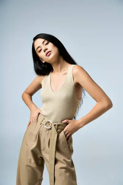 A young woman in a tan tank top and beige pants poses confidently against a light blue background. — Stock Photo