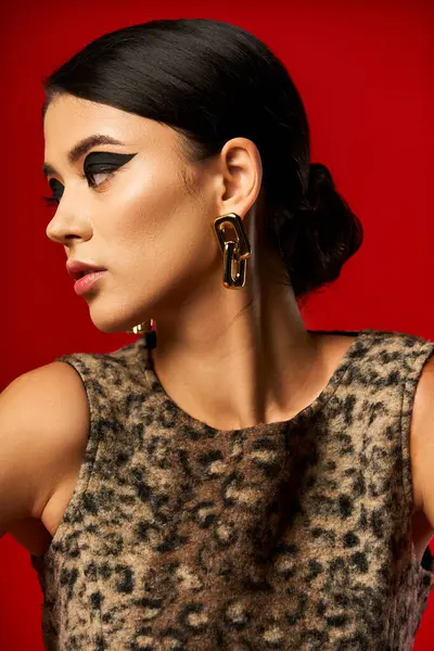 Dark haired woman with bold makeup poses against red backdrop in leopard-print dress. — Stock Photo