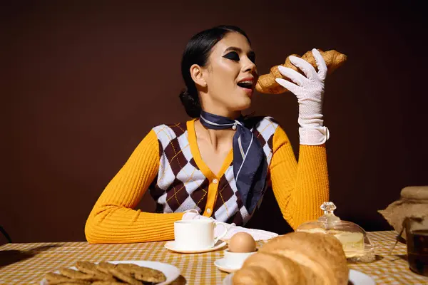 Eine junge Frau im gelben Pullover und weißen Handschuhen nimmt am Frühstückstisch ein Croissant.. — Stockfoto