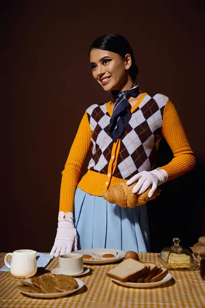 Uma jovem mulher em uma roupa elegante segura um croissant enquanto posando perto de uma mesa de café da manhã. — Fotografia de Stock