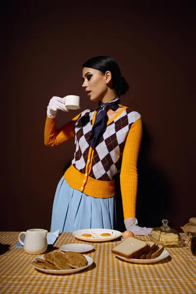 A young woman in a yellow and brown cardigan drinks coffee at a table set with breakfast. — Stock Photo