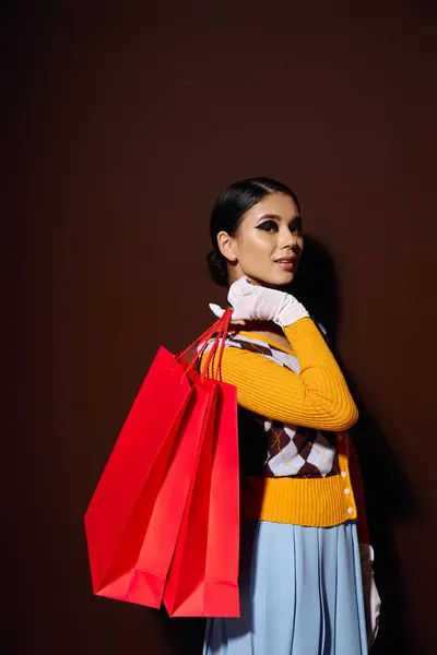 Uma jovem mulher em uma camisola amarela segura sacos de compras vermelhos na frente de um fundo marrom. — Fotografia de Stock