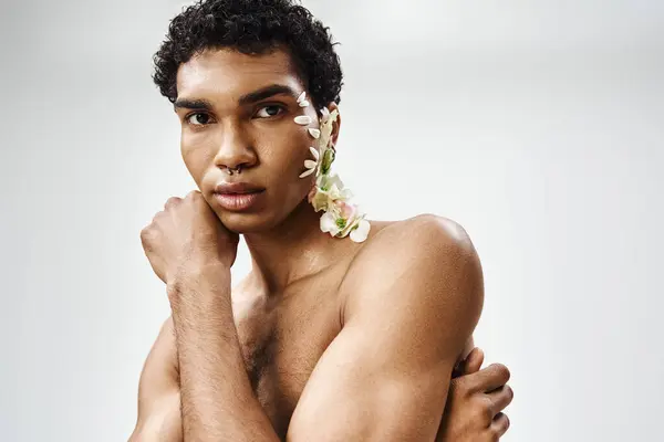 Un joven y musculoso afroamericano posa sobre un fondo gris, adornado con flores blancas en su rostro y cuerpo. — Stock Photo