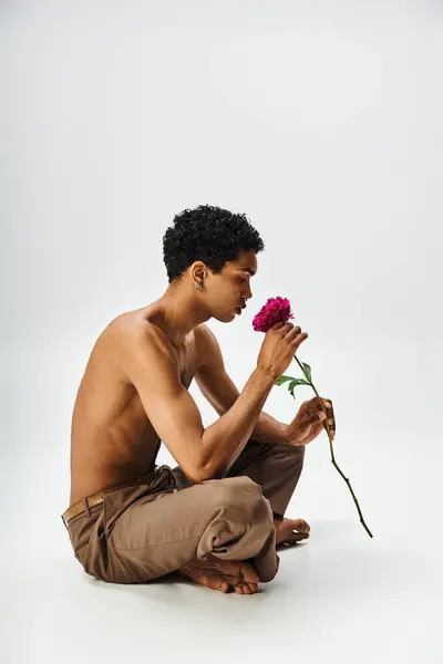 A young African American man smelling flower, sits on a grey background, showcasing beauty and diversity. — Stock Photo