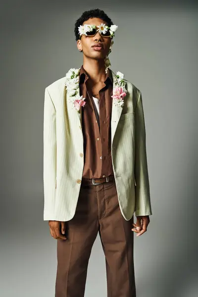 A young African American man poses in a stylish blazer, adorned with flowers around his neck and on his face, against a grey background. — Stock Photo