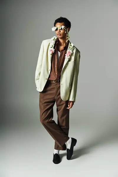 A young African American man poses with flowers adorning his neck and face, wearing a stylish blazer on a gray background. — Stock Photo