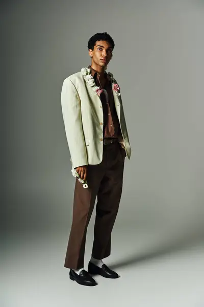 A confident young Black man in a stylish blazer, wearing flowers, stands out against a simple gray background, showcasing his beauty. — Stock Photo