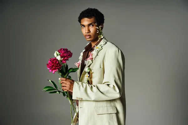 A young African American man in a stylish blazer poses with flowers adorning his neck and face against a grey backdrop. — Stock Photo
