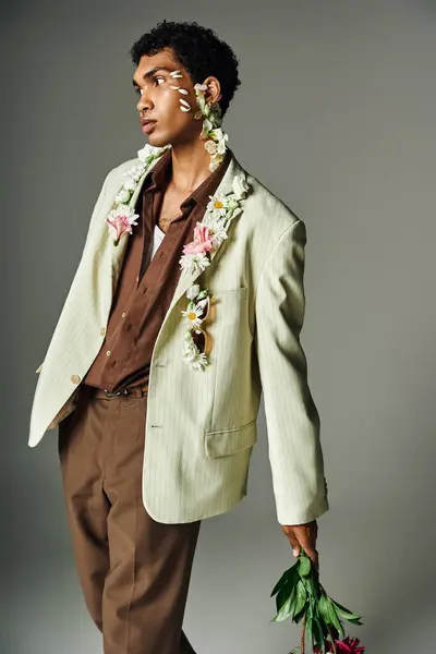 Un giovane afroamericano posa in un blazer elegante, ornato di fiori sul collo e sul viso, su uno sfondo grigio. — Foto stock