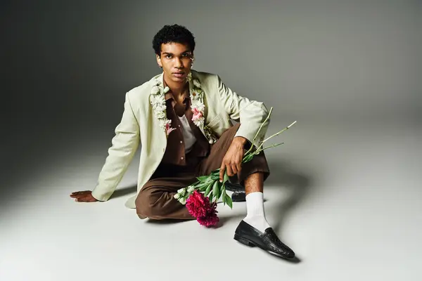 A young African American man in a stylish blazer sits with flowers around his neck against a grey background. — Stock Photo
