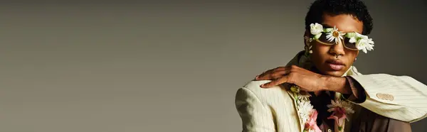 A young African American man wearing a stylish blazer poses against a grey background with flowers adorning his face and neck. — Stock Photo