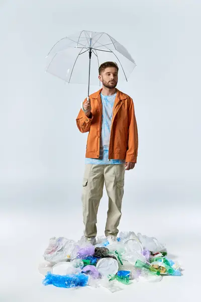 Ein Mann mit orangefarbener Jacke und durchsichtigem Regenschirm steht über einem großen Haufen Plastikmüll. — Stockfoto