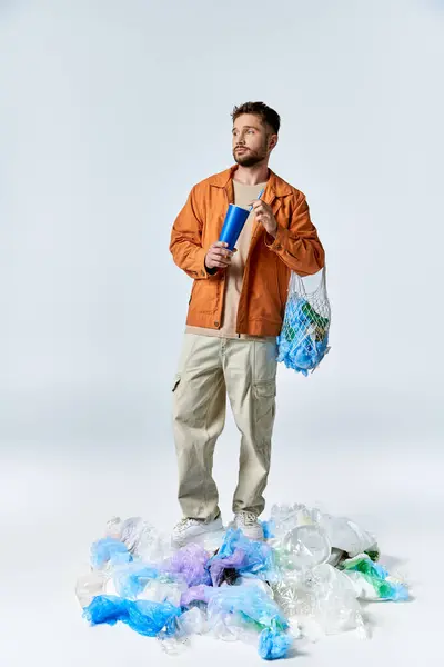 A man stands on a pile of plastic waste, holding a blue cup and a net bag filled with more plastic. — Stock Photo