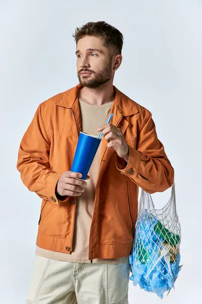 Un homme tient une tasse bleue et une paille tout en portant un sac de filet rempli de déchets plastiques. — Photo de stock