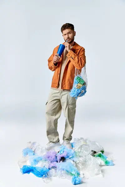 A man stands on a large pile of plastic waste, holding a blue cup and a mesh bag. — Stock Photo