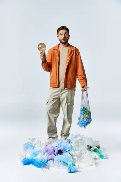 Мужчина стоит на насыпи пластиковых отходов, держа в руках лампочку и сетку мешка. — стоковое фото