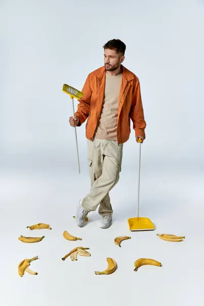 A man stands in a white studio, surrounded by banana peels, holding a broom and dustpan. — Stock Photo