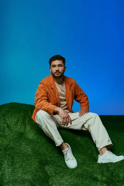 Un homme vêtu d'une veste orange est assis sur une colline verte avec un fond bleu. — Photo de stock