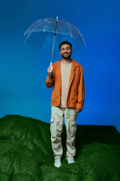 A man stands on a green hill, holding a clear umbrella, smiling at the camera. — Stock Photo