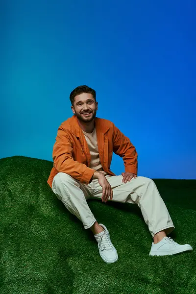A man in an orange jacket and white pants sits on a patch of green grass. — Stock Photo