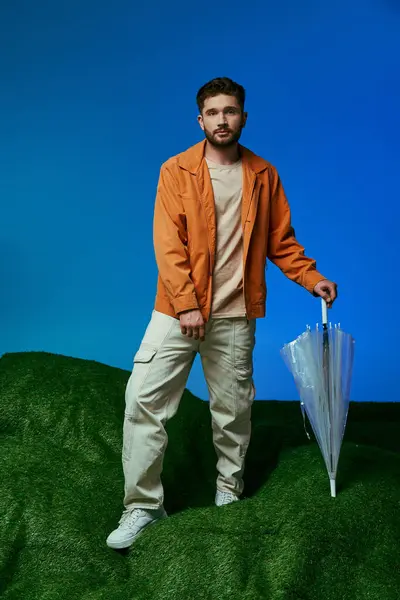 A man in an orange jacket stands on a green hill, holding a transparent umbrella. — Stock Photo