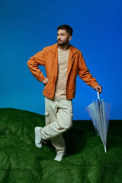 Homme en veste orange pose sur la colline avec parapluie, fond bleu. — Photo de stock