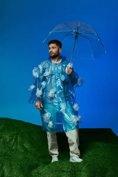 Um homem está em uma colina gramada, vestindo uma capa de chuva feita de sacos de plástico e segurando um guarda-chuva claro. — Fotografia de Stock
